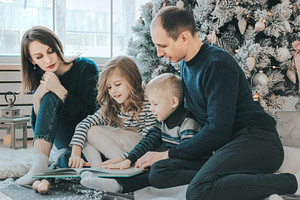 girl and boy reading book sitting between man and woman beside Christmas tree
